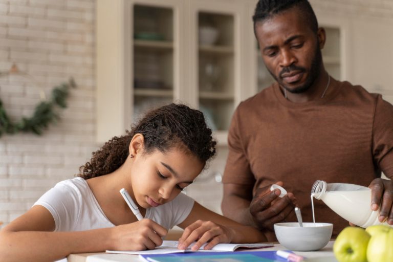 A Black father pours milk while his daughter writes at the kitchen table, representing the importance of family presence in youth suicide prevention.